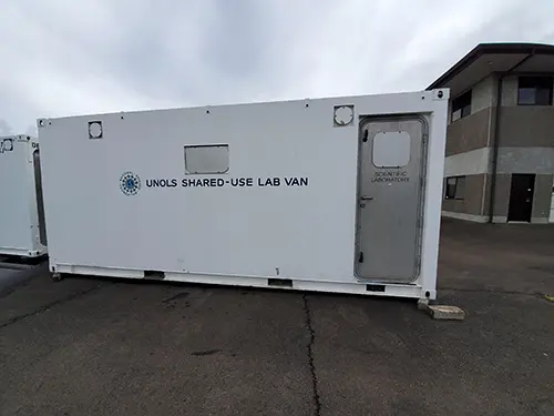 The white exterior of a portable lab van in front of a building.