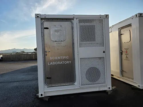 The white exterior of the end of a portable lab van showing a silver door.