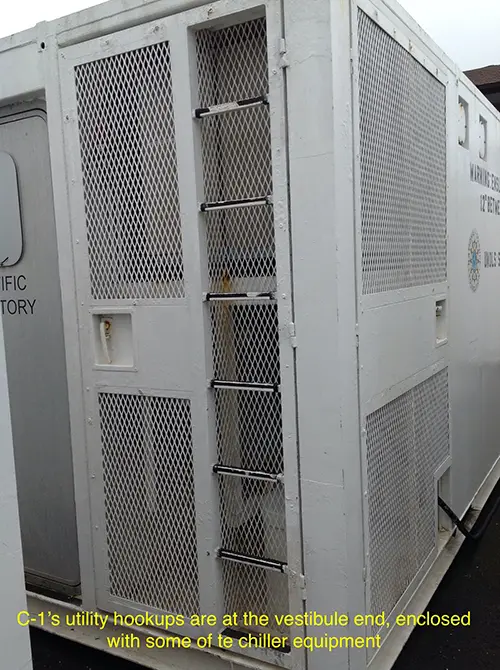 The white exterior of a lab van with a silver entry door and utility hookups enclosed with chiller equipment.