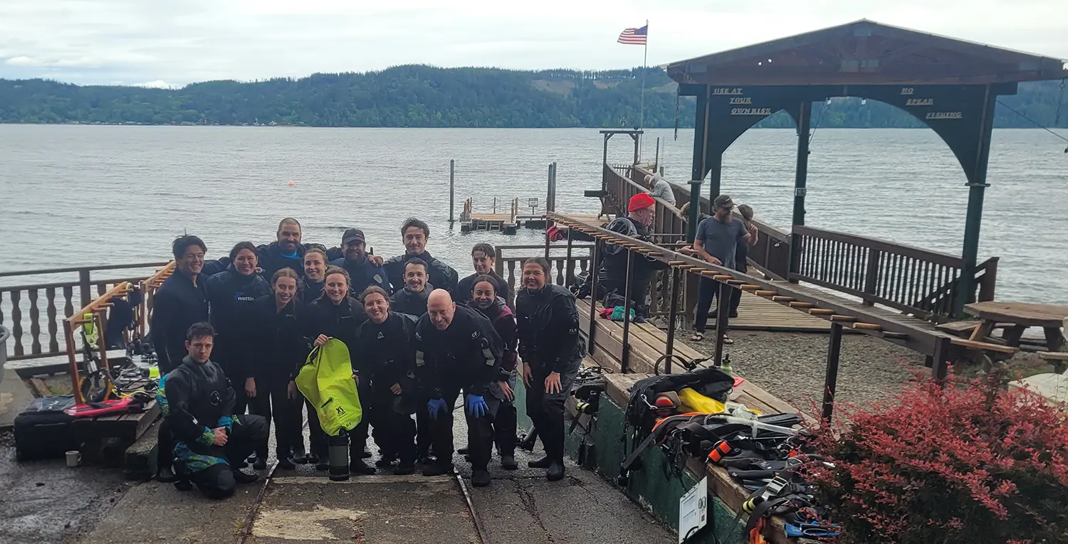 A group of people wearing scuba suits gather for a photo at the waters edge.