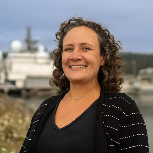 A woman with curly hair smiles, a large boat is in the background.