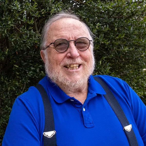 An older man wearing tinted round glasses, suspenders and a blue shirt.