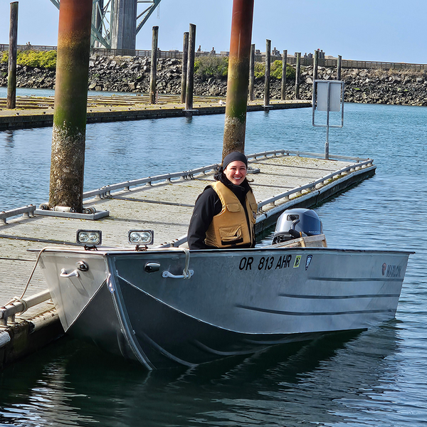 A person in a lifevest sits in an open metal boat tied up at a dock.